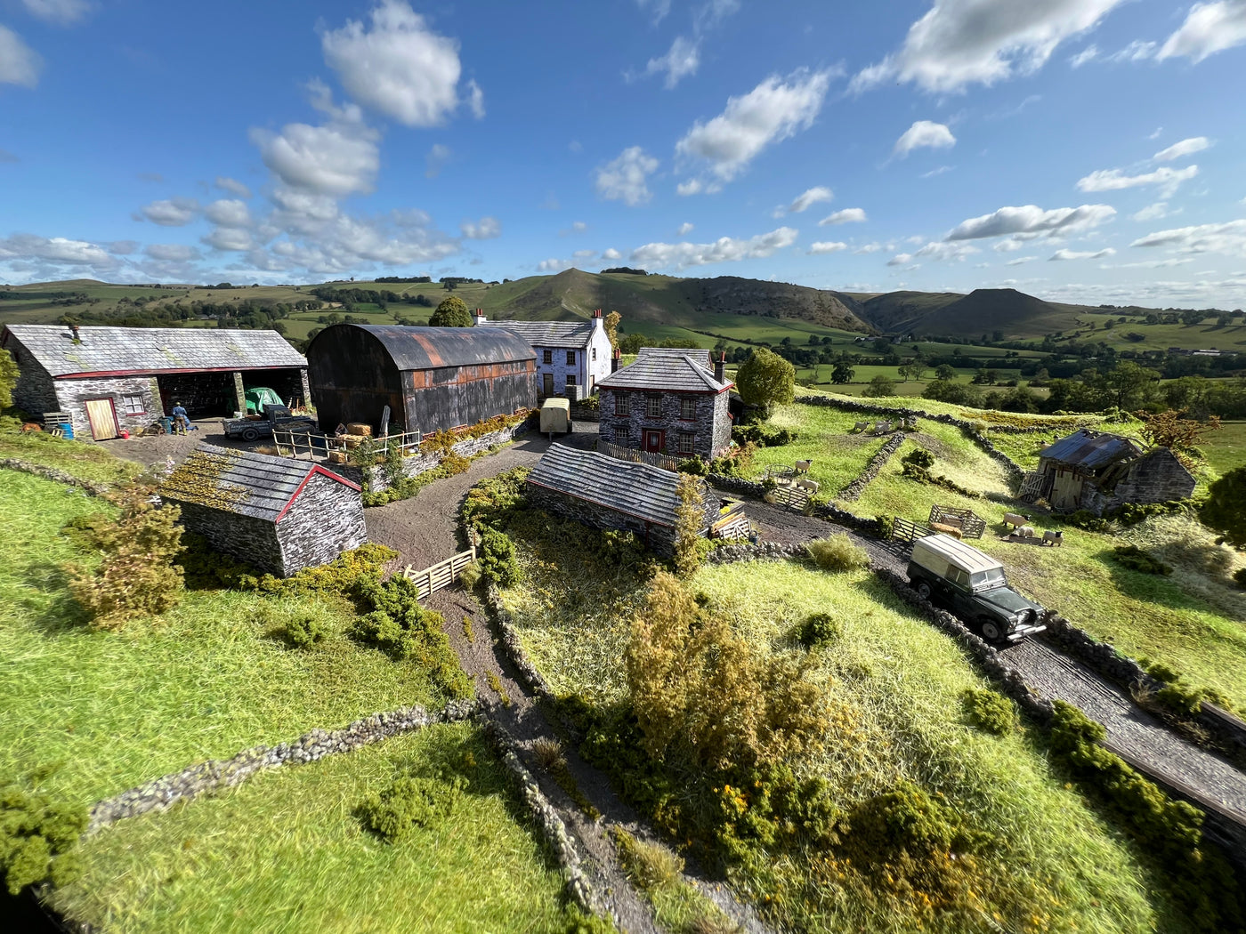 A miniature OO gauge model diorama of a rural village scene, featuring stone cottages, a large barn, a vintage white van on a road, and a model car, all set against a realistic green hillside background under a blue sky with clouds.
