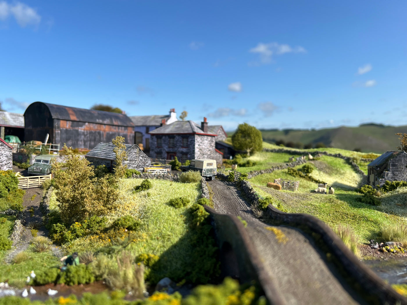 A miniature OO scale model village scene with stone buildings, a curved road, grassy fields, shrubs, and a blue sky in the background.